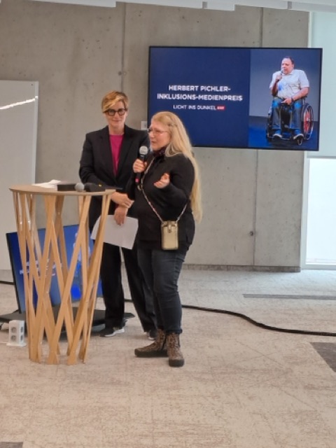 Zwei Frauen am Rednerpult, eine spricht ins Mikrofon. Im Hintergrund: Bildschirm mit dem Titel des Herbert Pichler Inklusions-Medienpreises.