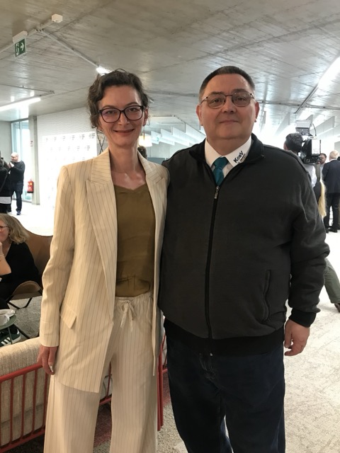 Mag.a Viktoria Antrey (KOBV Kommunikation) und KOBV-Vizepräsident Georg Fitzthum stehen nah beieinander und lächeln in die Kamera, im Hintergrund eine moderne Betonarchitektur und Gäste in Gesprächssituationen.
