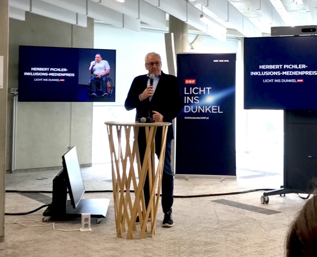 Prof. Pius Strobl spricht am Rednerpult, umgeben von Bildschirmen mit dem Titel „Herbert Pichler Inklusions-Medienpreis“ und einem Porträt eines Mannes im Rollstuhl.