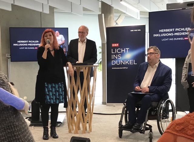 Eine Frau mit rotem Haar spricht ins Mikrofon, neben ihr ein Mann im Rollstuhl, dahinter Bildschirme und ein Roll-Up mit dem Schriftzug „Licht ins Dunkel“.