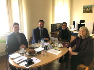 Zwei Männer und zwei Frauen sitzen mit Laptop, Getränken, Kuchen und Unterlagen an einem Tisch in einem Büro.