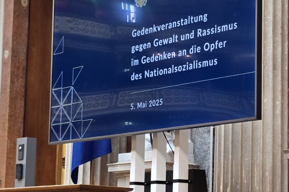 Ein Rednerpult aus Holz und daneben ein großer Bildschirm, auf dem der Titel der Gedenkveranstaltung zu lesen ist.