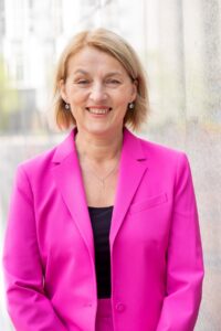 Eine Frau mit kinnlangem blondem Haar in pinkfarbenem Blazer lächelt freundlich geradeaus ins Bild.