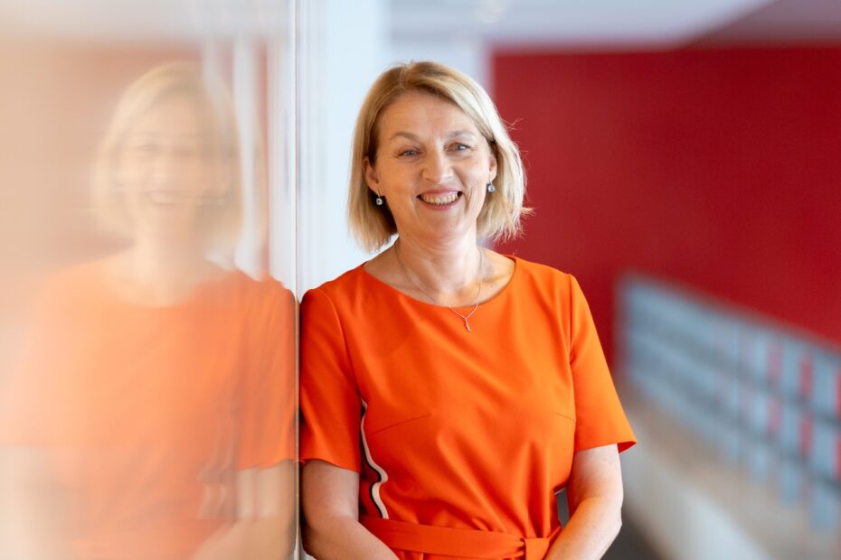 Eine Frau mit kinnlangem blondem Haar in orangenem Kleid lehnt an einer spiegelnden Fläche, die sie reflektiert, und lächelt dabei freundlich.