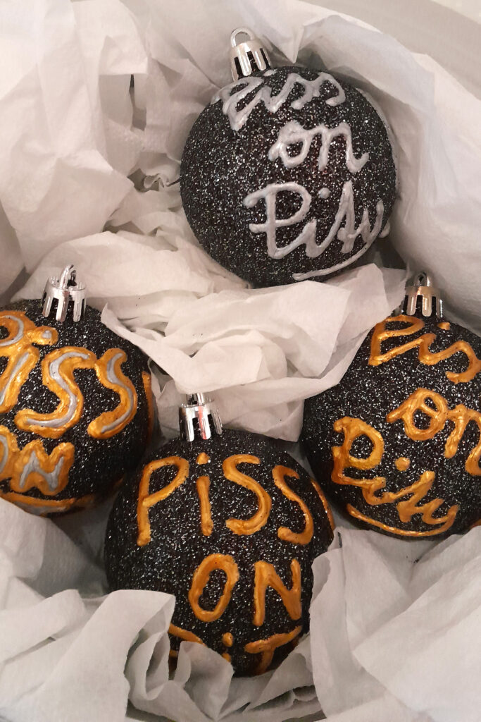 Vier schwarz glitzernde Christbaumkugeln liegen in einem Korb. Sie haben die Aufschrift "Piss on Pity".