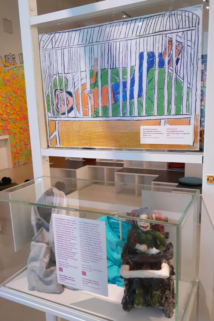 Über einem Schaukasten, der eine Figur, die eine Rollstuhlfahrerin darstellt, enthält, hängt eine Zeichnung von einer jungen Frau in einem Gitterbett.