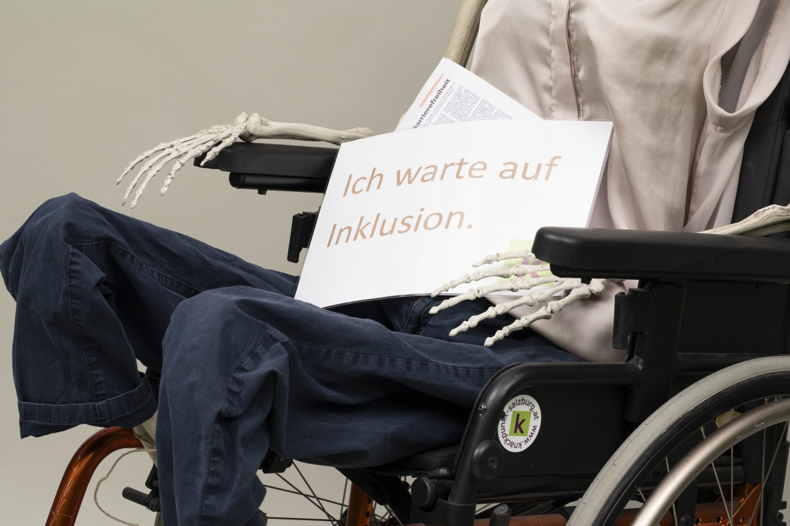 Ein angezogenes Skelett sitzt in einem Rollstuhl und hält ein Schild mit der Aufschrift "Ich warte auf Inklusion." in den Händen