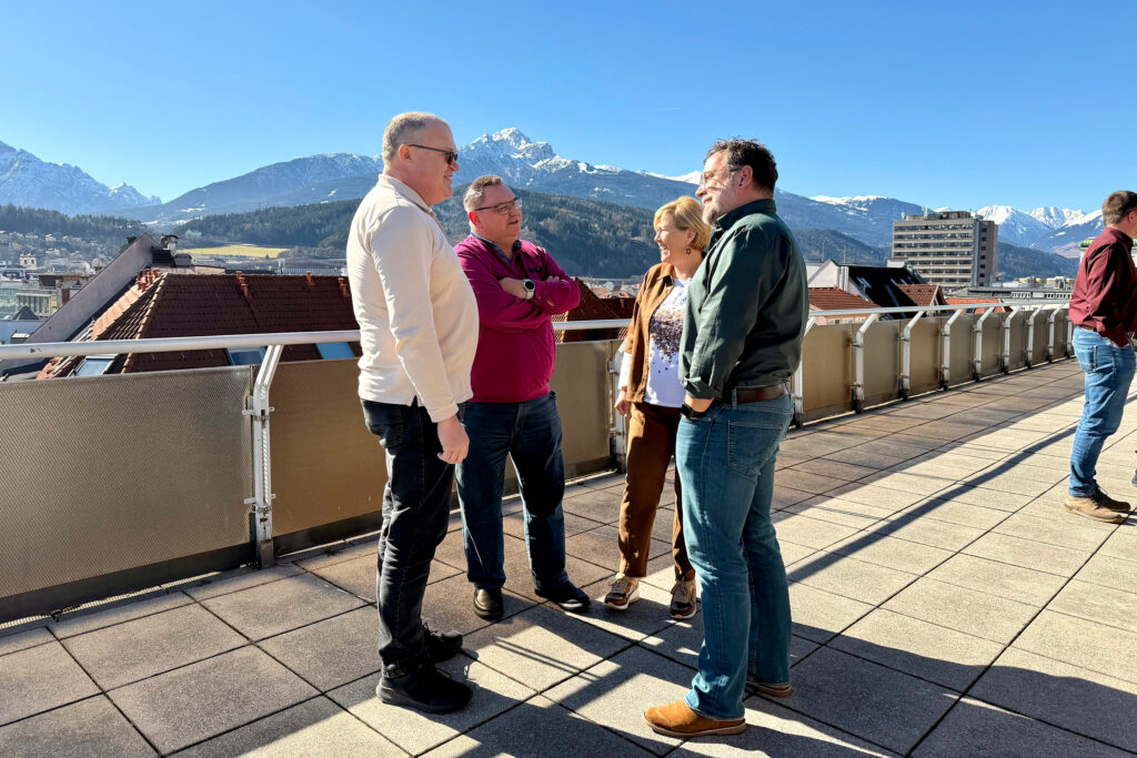 Vier Menschen unterhalten sich auf der Terrasse eines Gebäudes, im Hintergrund sind die Tiroler Berge zu sehen
