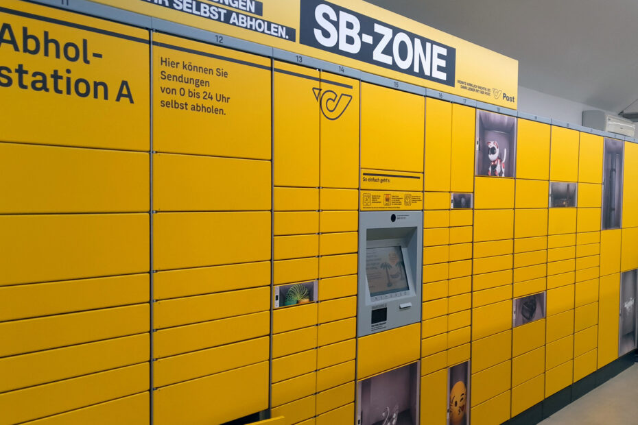 Abholstation Post-Abholstation mit zahlreichen Fächern auf unterschiedlichen Höhen und in unterschiedlichen Größen. In der Mitte befindet sich ein Bildschirm zum Bedienen, die Fächer sind in Gelb gehalten.
